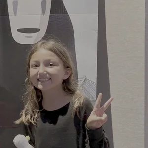 Jojo smiling and holding up a peace sign, standing in front of a big painting of No-Face from Spirited Away.